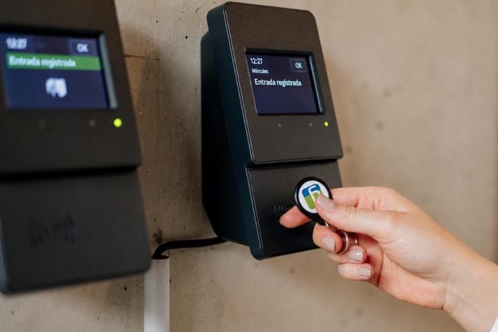 Una persona acerca una ficha de identificación a un terminal digital de control horario. En la pantalla aparece el mensaje «Entrada registrada»; a la izquierda se ve un segundo terminal similar.