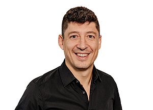 Porträt eines lächelnden Mannes mit kurzen dunklen Haaren und schwarzem Hemd vor weißem Hintergrund.